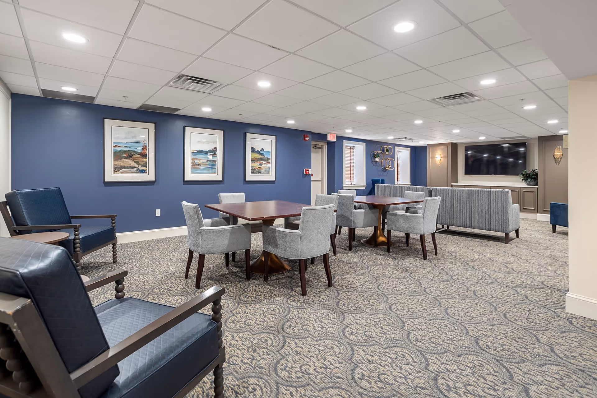A spacious common area with patterned carpet, blue accent wall with three framed landscape paintings, two wooden tables surrounded by gray upholstered chairs, blue armchairs, and a large flat-screen TV mounted on a beige wall with built-in cabinetry below.
