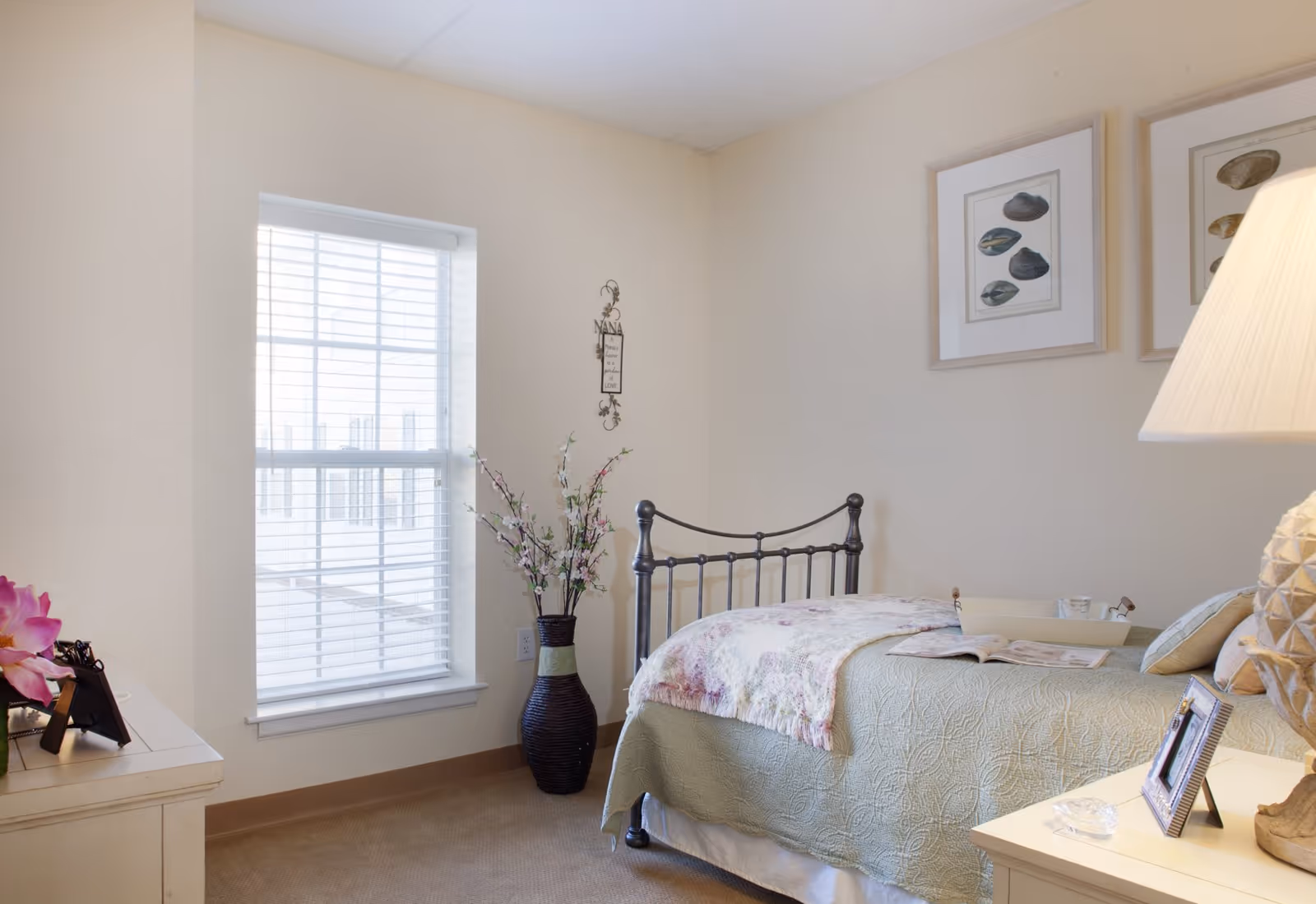 A cozy bedroom with a metal bed frame, a green quilted bedspread, and a floral throw blanket. There is a window with white blinds letting in natural light, a large vase with decorative branches, framed shell artwork on the wall, and a bedside table with a lamp, picture frame, and small decorative items.