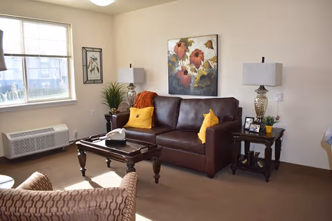 A cozy living room with a brown leather sofa adorned with yellow pillows and an orange throw blanket. There are two matching side tables with decorative lamps and small plants. A coffee table with a tissue box sits in front of the sofa. The room has beige walls with floral artwork and a window letting in natural light.