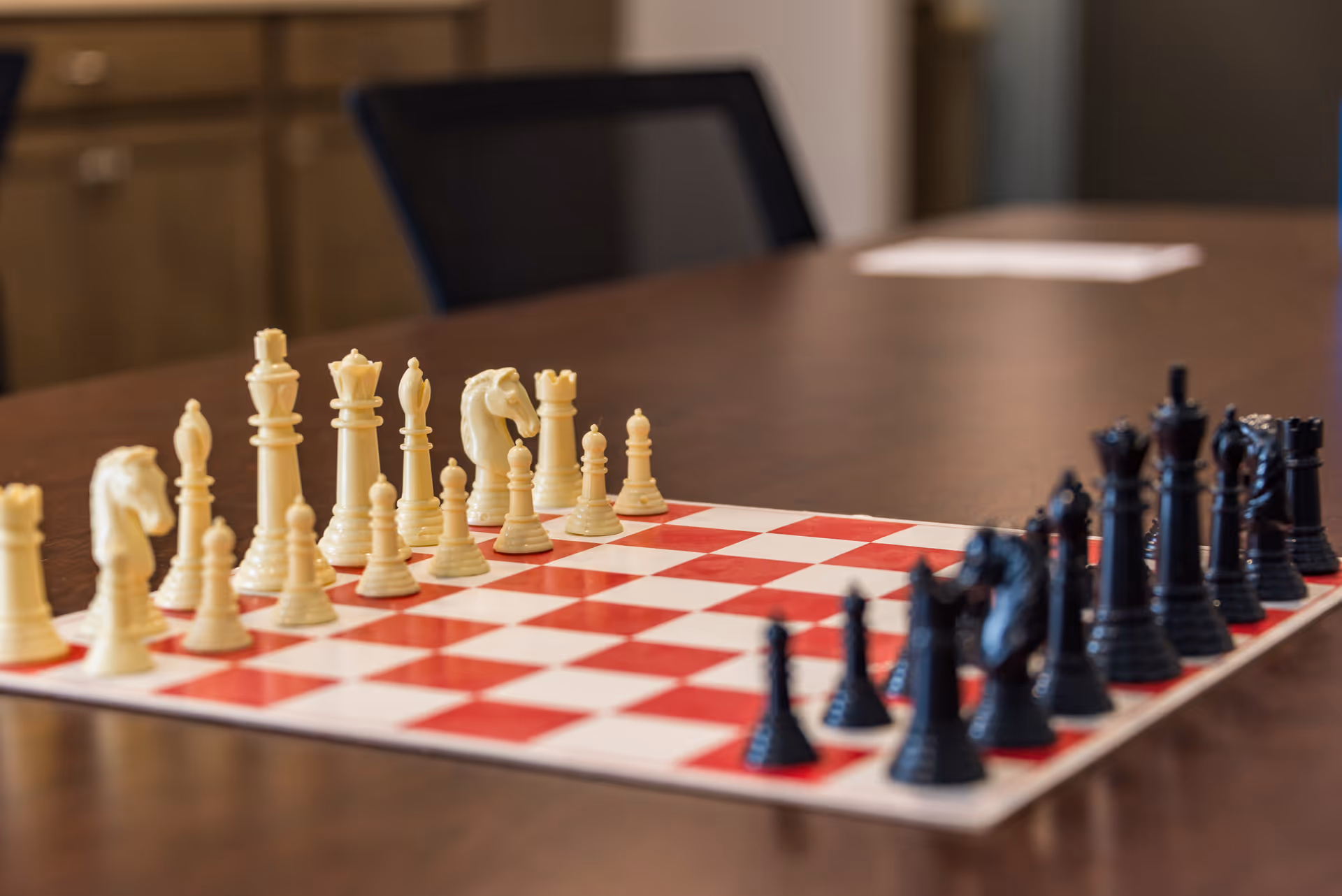 A chessboard with white and black chess pieces set up for a game on a wooden table in a room with cabinets and a chair in the background.