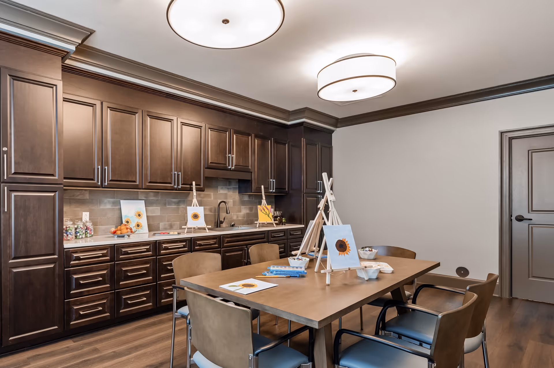 A well-lit room with dark wooden cabinets along one wall and a wooden table surrounded by six chairs. The table has several small easels with sunflower paintings, paint palettes, and art supplies, suggesting an art activity space. The room has two modern ceiling lights and a closed door on the right side.