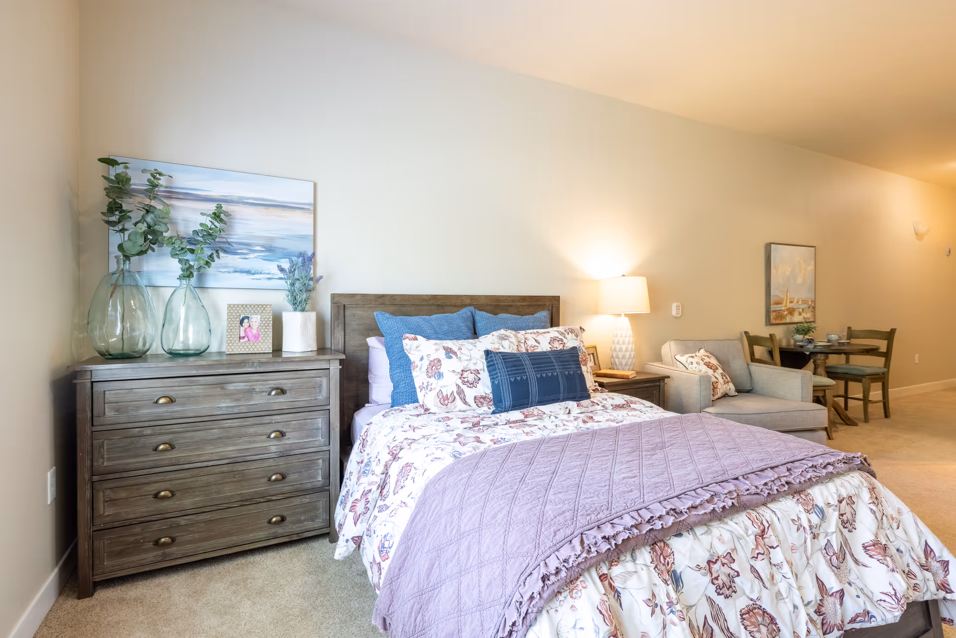 Bright furnished bedroom with a bed, wooden dresser, nightstand lamp, and a nearby seating and dining area.