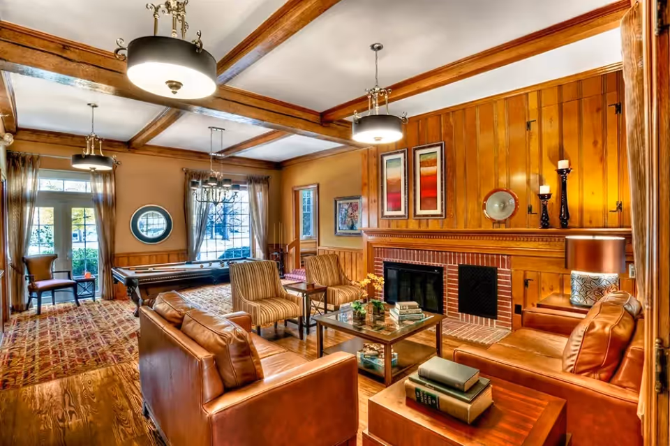 A cozy living room with wooden paneling and exposed ceiling beams, featuring two leather sofas, two striped armchairs, a glass coffee table with books and flowers, a brick fireplace with candles and framed artwork above it, a pool table near large windows with sheer curtains, and warm lighting from ceiling fixtures and a table lamp.