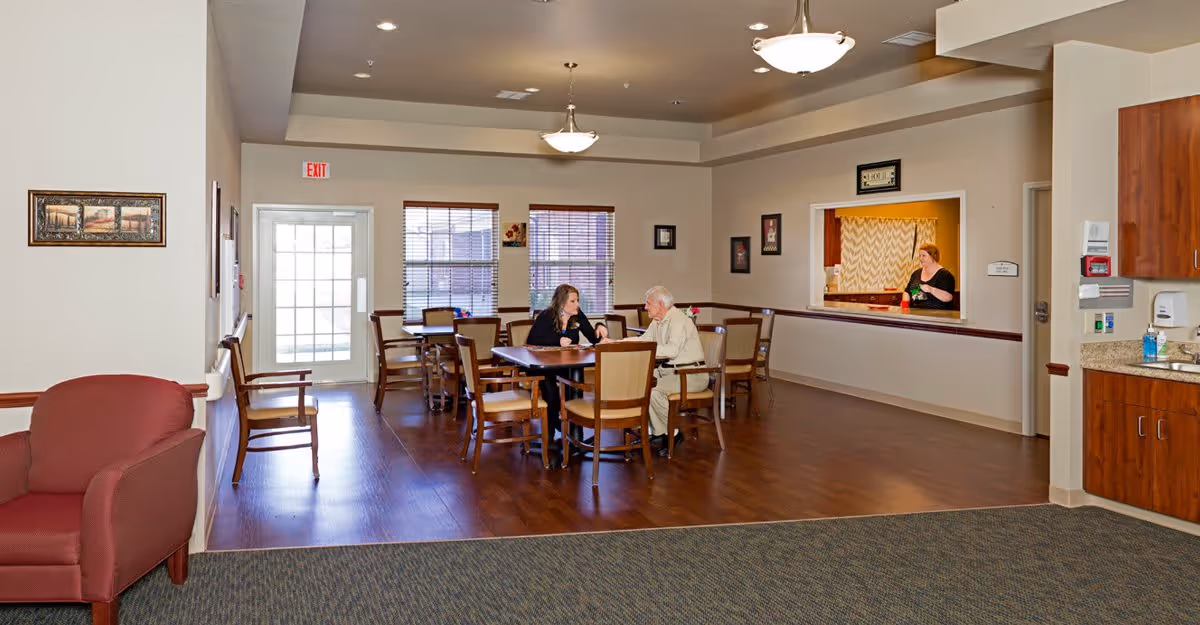 A dining area in a senior living facility with wooden tables and chairs. Two people are seated at a table having a conversation. A woman is seen behind a serving window in the background. The room has large windows with blinds, wooden flooring, and beige walls. There is a red armchair and additional chairs along the left wall.
