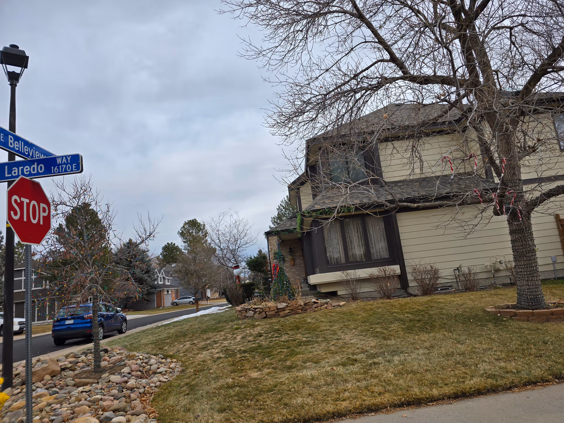 Street corner with a stop sign and street signs for Laredo Way and Belleview. A residential house with Christmas decorations including candy canes on a tree and a decorated tree in the yard is visible. The sky is cloudy and the trees are bare.