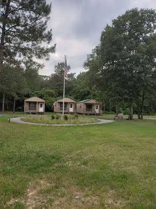 A grassy outdoor area with a circular paved path surrounding a flagpole flying the American flag. Three small beige cabins are visible in the background, surrounded by tall green trees under a cloudy sky.