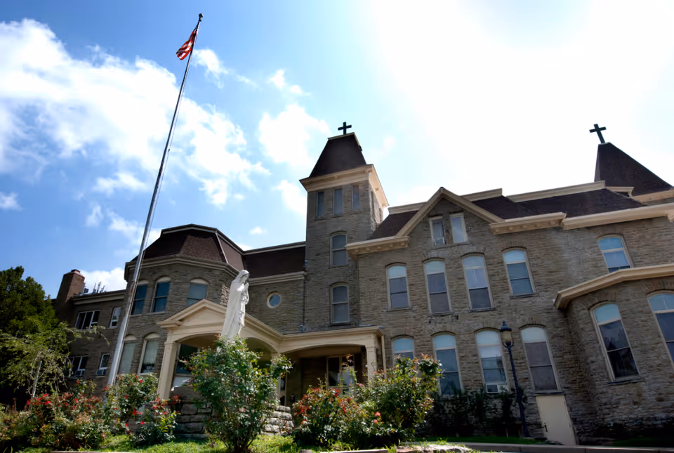 Exterior view of a large stone building with multiple windows and two towers topped with crosses. There is a statue in front of the building surrounded by bushes and flowers, and an American flag on a tall flagpole against a partly cloudy sky.