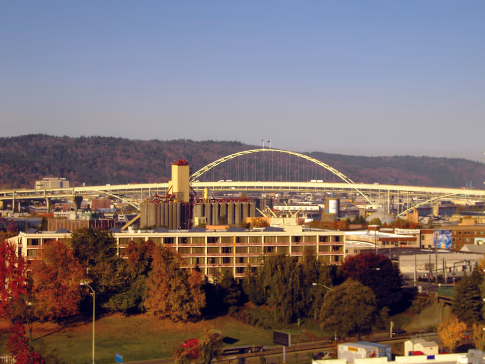 Wide exterior city view showing an arched bridge over industrial buildings with trees in the foreground.