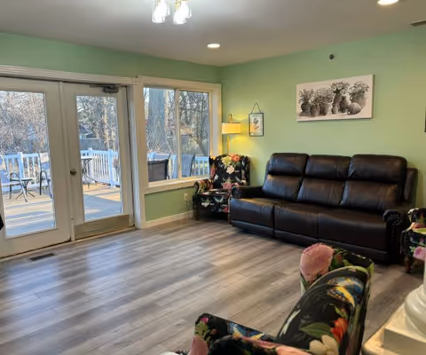 A cozy living room with light green walls, a dark brown leather sofa, and two floral-patterned armchairs. There is a floor lamp and a framed picture of potted plants on the wall. Large windows and glass doors lead to an outdoor deck with patio furniture, allowing natural light to fill the room. The floor is light-colored wood.