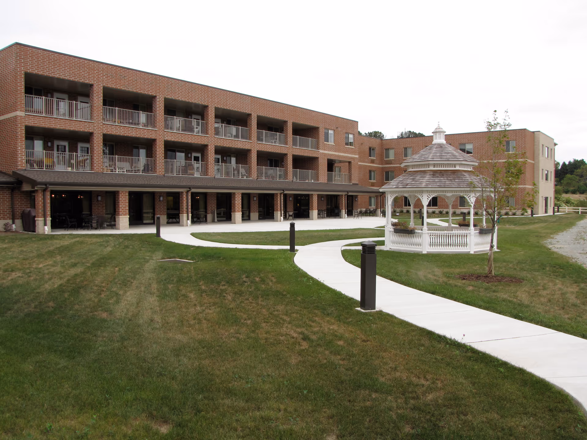 Brick three-story senior living building with balconies, a white gazebo and a curved sidewalk across a grassy courtyard.