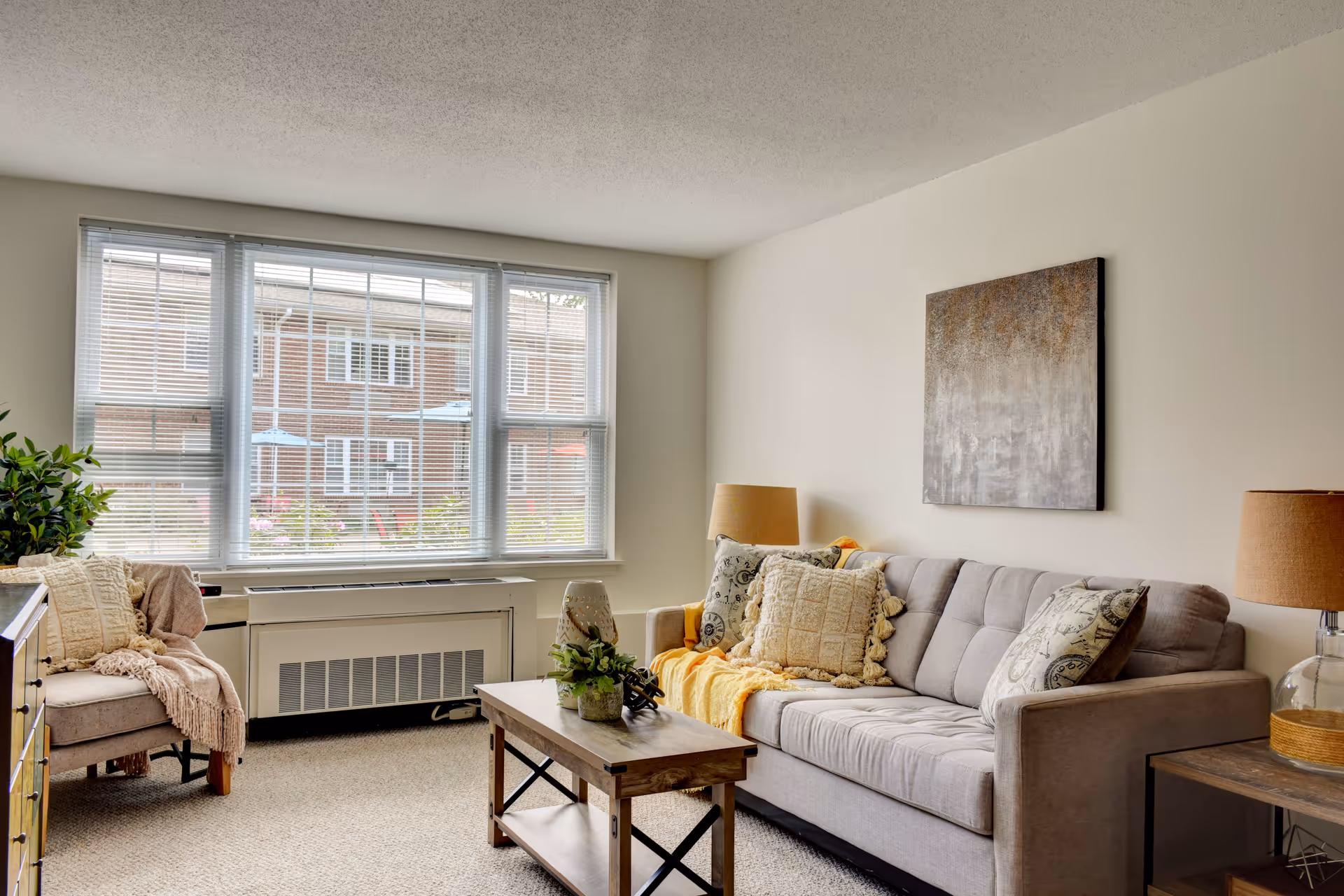 Bright living room with a gray sofa, wooden coffee table, armchair by a large window with blinds, and decorative pillows and lamps.