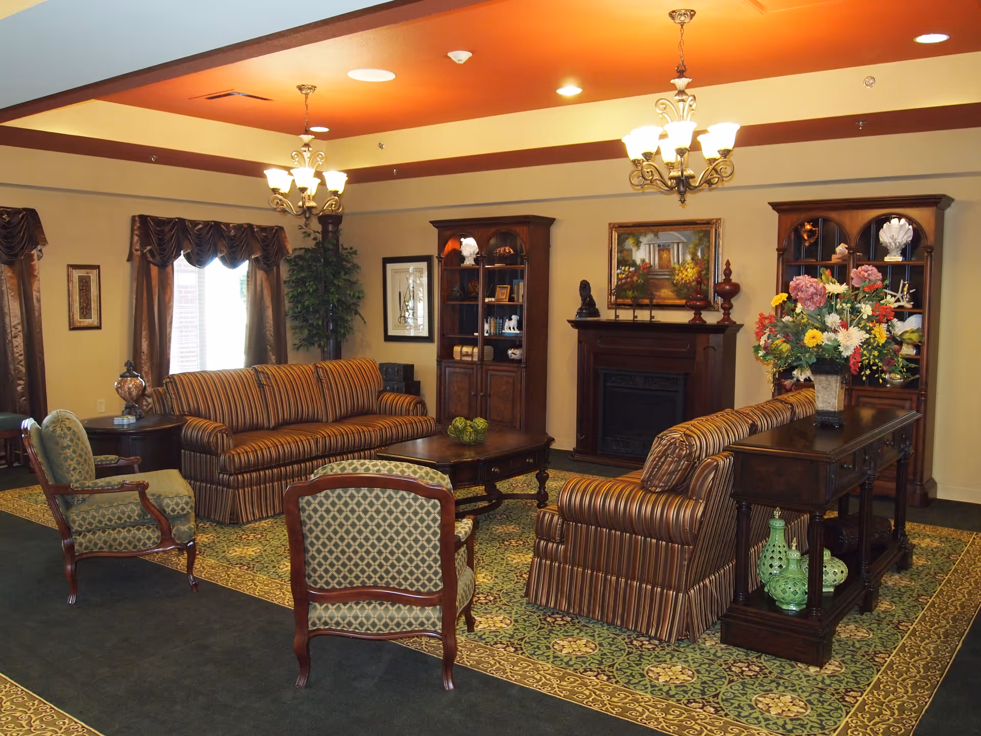 A warmly furnished communal living room with striped sofas, upholstered chairs, bookshelves, a fireplace, and a floral arrangement on a console table.