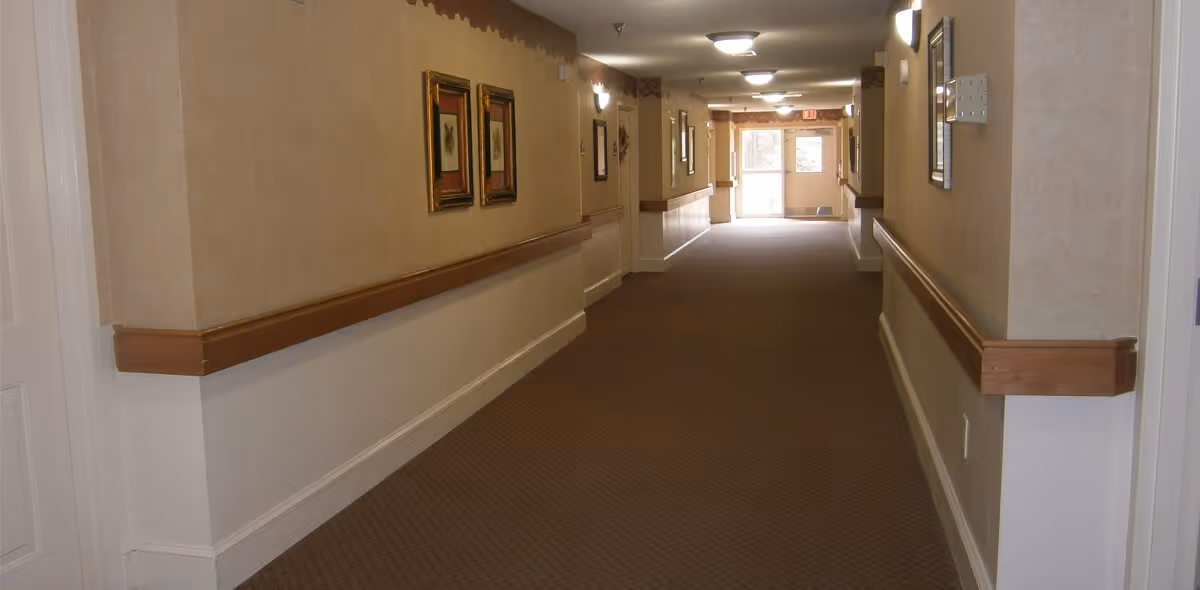 A long, carpeted hallway in a senior living facility with beige walls, wooden handrails on both sides, framed artwork hanging on the walls, and ceiling lights. At the end of the hallway, there is a glass door letting in natural light.