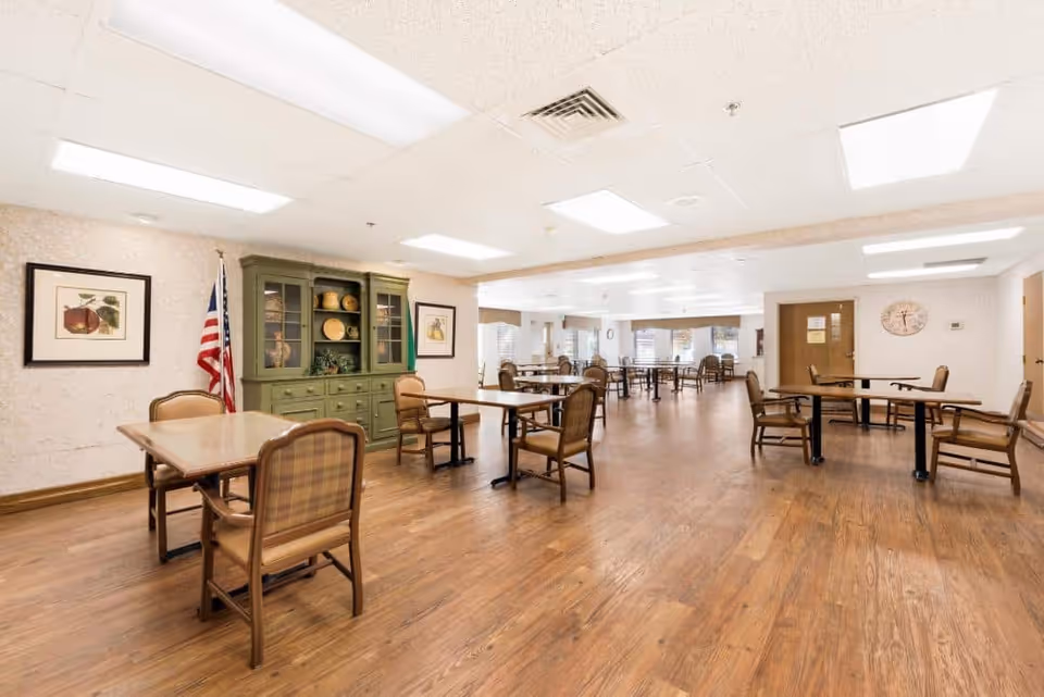 A spacious dining room with multiple wooden tables and cushioned chairs arranged neatly. The room has a wooden floor, white ceiling with fluorescent lights, and walls decorated with framed pictures and an American flag. A green cabinet with decorative items is against one wall.