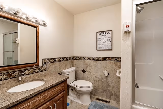 Clean bathroom with a vanity and mirror on the left, a toilet in the center, tiled walls and a shower stall on the right.