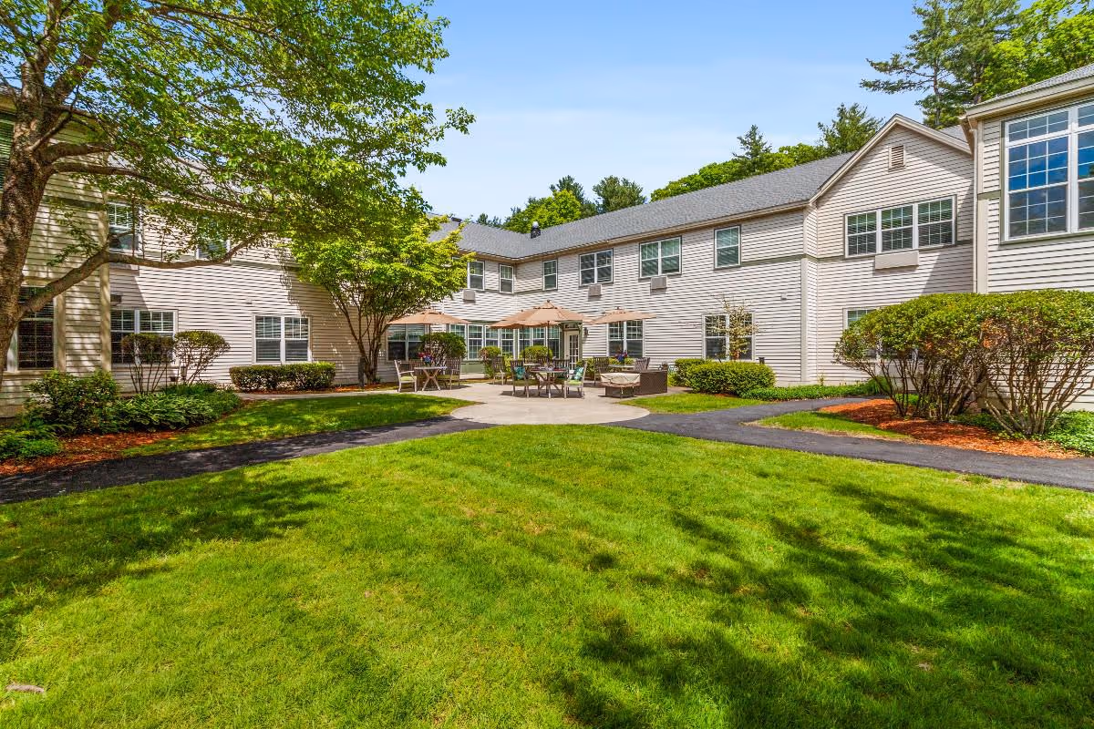 Outdoor courtyard area of Benchmark at Tatnuck Park featuring a well-maintained green lawn, paved walkways, patio furniture with umbrellas, and surrounding two-story building with multiple windows under a clear blue sky.