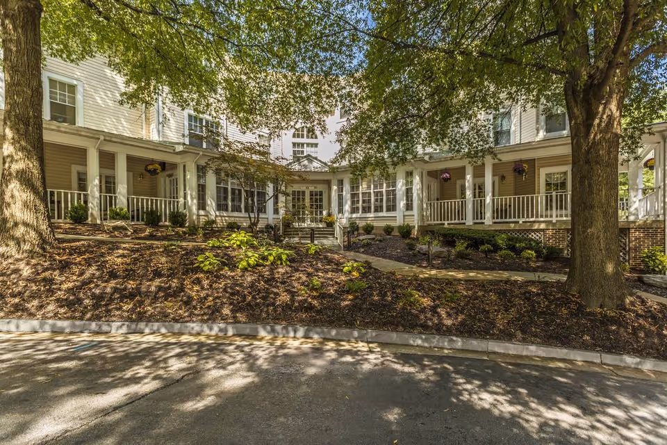 Front exterior of a multi-story senior living building with white columns, a covered wraparound porch, mature trees, and landscaped beds.