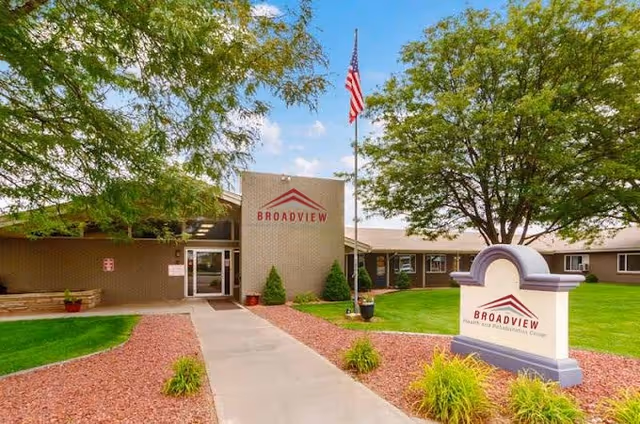 Exterior view of Broadview Health and Rehabilitation facility showing the entrance with a walkway, green lawn, trees, an American flag on a flagpole, and a sign with the facility name.