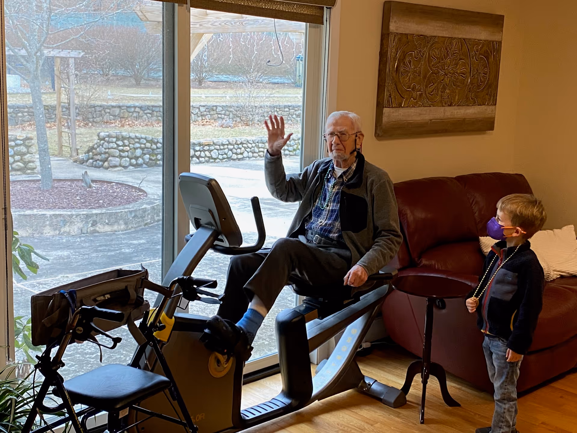 An elderly man is sitting on a stationary exercise bike indoors near a large window, waving his hand. A young boy wearing a purple face mask and a beaded necklace stands nearby, looking at the man. There is a red leather couch and a decorative wall hanging in the background, and a walker is positioned next to the exercise bike.