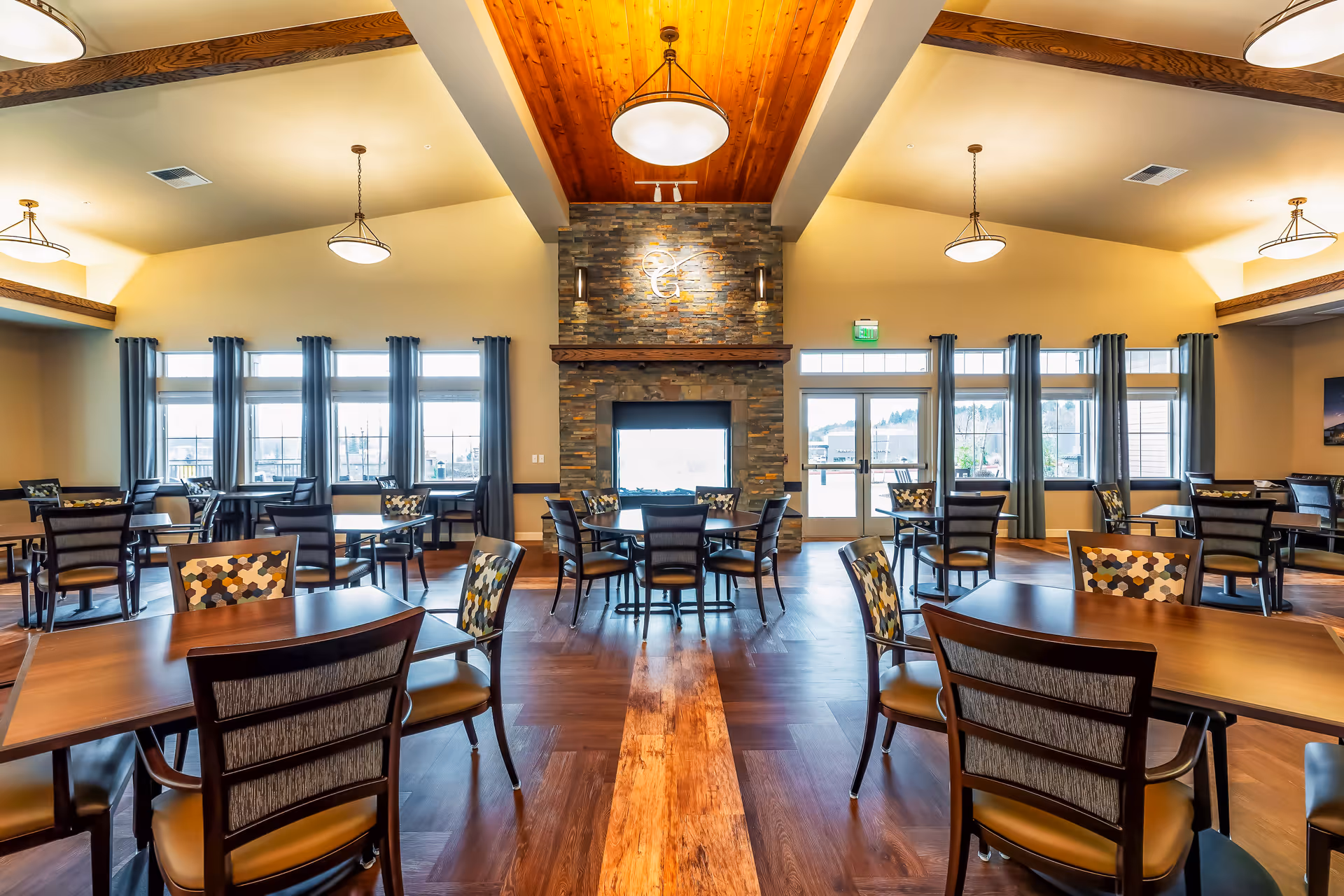 A spacious dining room with multiple wooden tables and chairs arranged neatly. The room features large windows with blue curtains allowing natural light to fill the space. A stone fireplace with a wooden mantel is centered on the far wall, topped with a decorative light fixture. The ceiling has wooden beams and hanging light fixtures, and the floor is made of polished wood.