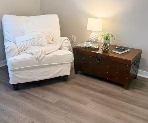 A cozy corner featuring a white cushioned armchair with a textured white throw blanket and pillow, next to a dark wooden chest used as a side table. On the chest are a small table lamp with a white shade, a potted plant, a pair of glasses, a book, and a decorative tray with a cup and saucer. The setting is on a light wood floor against a plain light-colored wall.