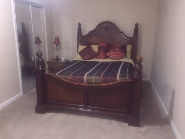 A bedroom with a large wooden bed featuring an ornate headboard and footboard. The bed is made with a striped bedspread and several decorative pillows in shades of red and gold. There are two matching bedside tables, each with a red lamp. A mirrored closet door is visible on the left side of the room. The walls are painted a light beige color and the floor is carpeted.