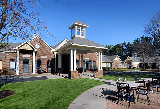 Exterior view of Belmont Village Senior Living Johns Creek featuring a brick building with a covered entrance supported by white columns, surrounded by green lawns and outdoor seating with tables and chairs under a clear blue sky.