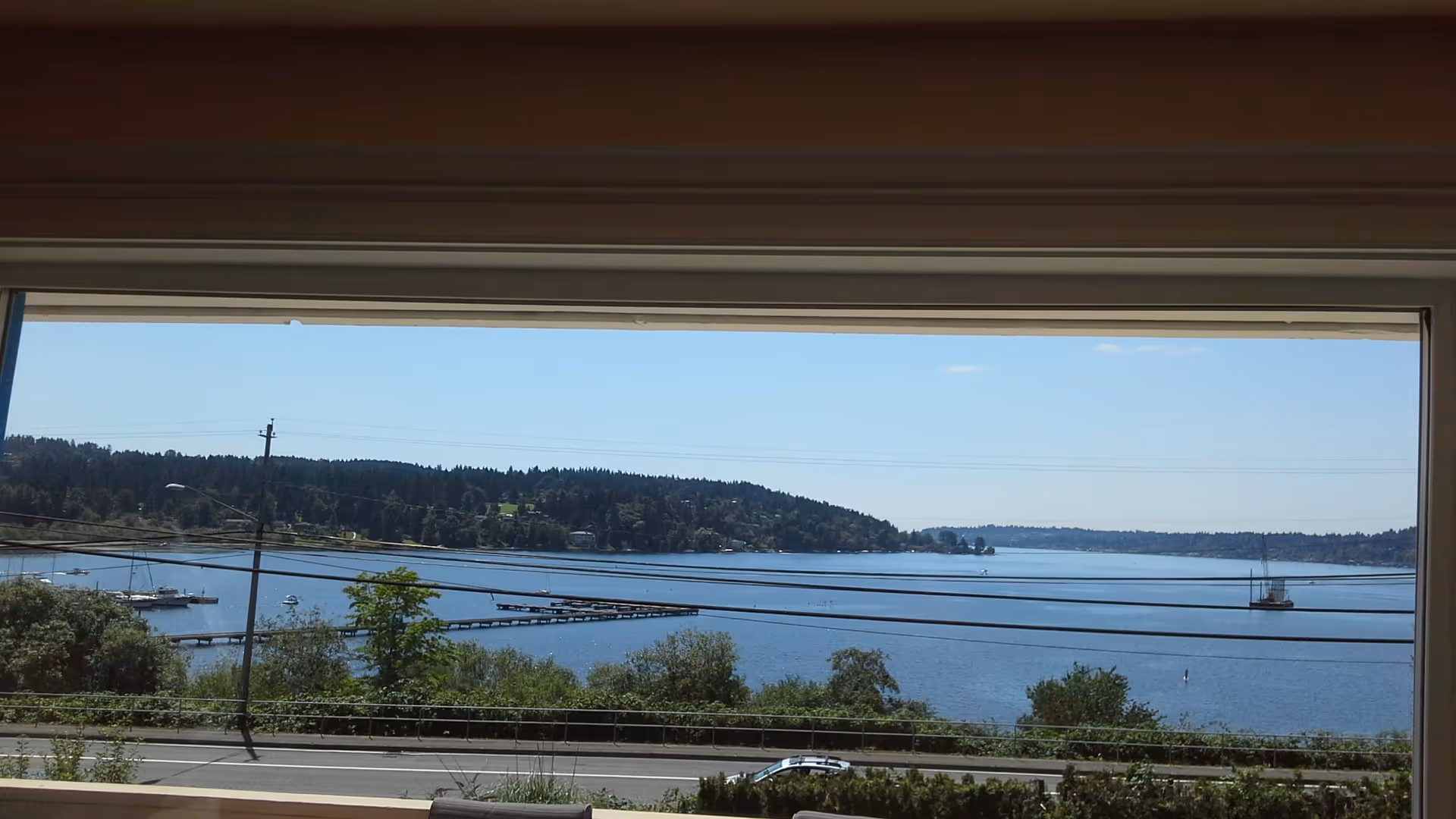 View through a large window showing a serene lake with docks, boats, and a tree-covered shoreline under a clear blue sky. A road with a car and power lines are visible in the foreground.