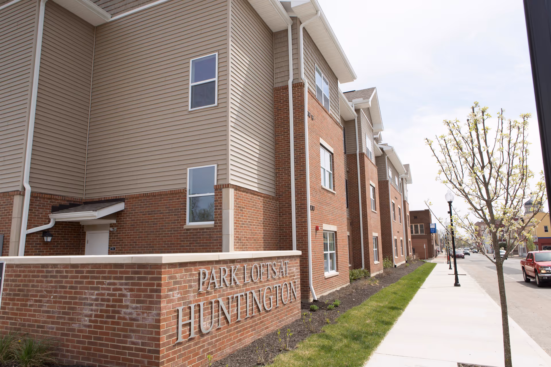 Exterior view of a multi-story residential building with brick and beige siding. A brick sign in front reads 'Park Lofts at Huntington'. There is a sidewalk, a small tree, and a street with parked cars visible.