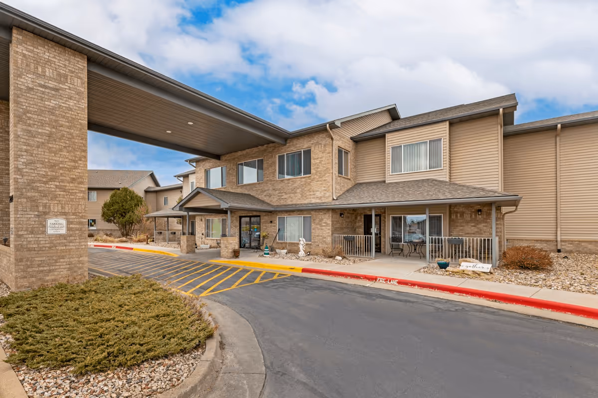 Exterior view of Arrowhead Lodge Senior Living facility showing a two-story building with a covered entrance, brick and siding facade, windows with blinds, a small patio area with a table and chairs, landscaped bushes, and a driveway with yellow and red painted curbs.