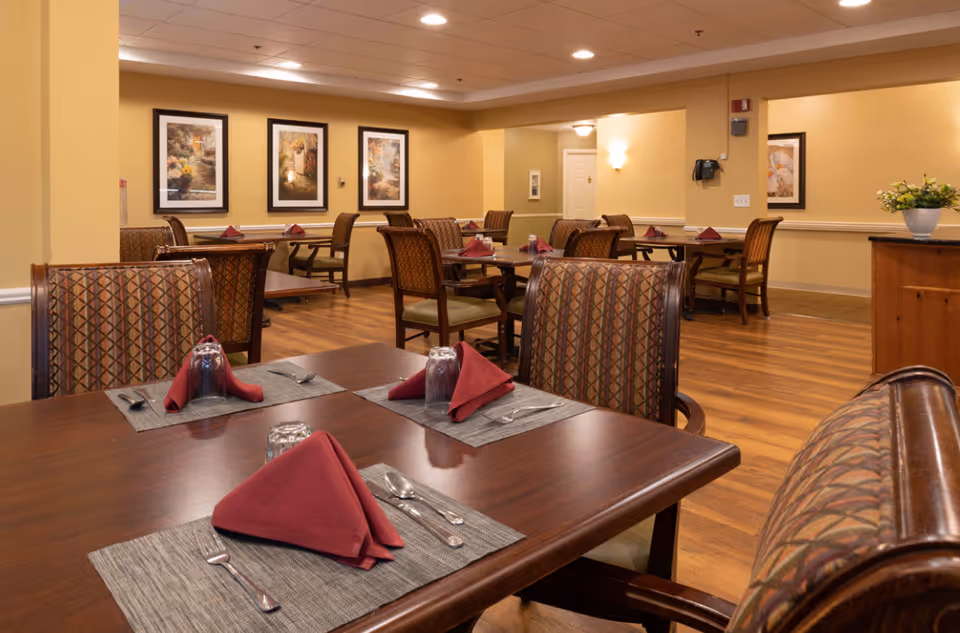 A dining room in a senior living facility with several wooden tables and cushioned chairs arranged neatly. Each table is set with gray placemats, silverware, upside-down glasses, and folded red napkins. The room has warm yellow walls adorned with framed artwork and features wood flooring. Soft lighting illuminates the space, creating a cozy and inviting atmosphere.
