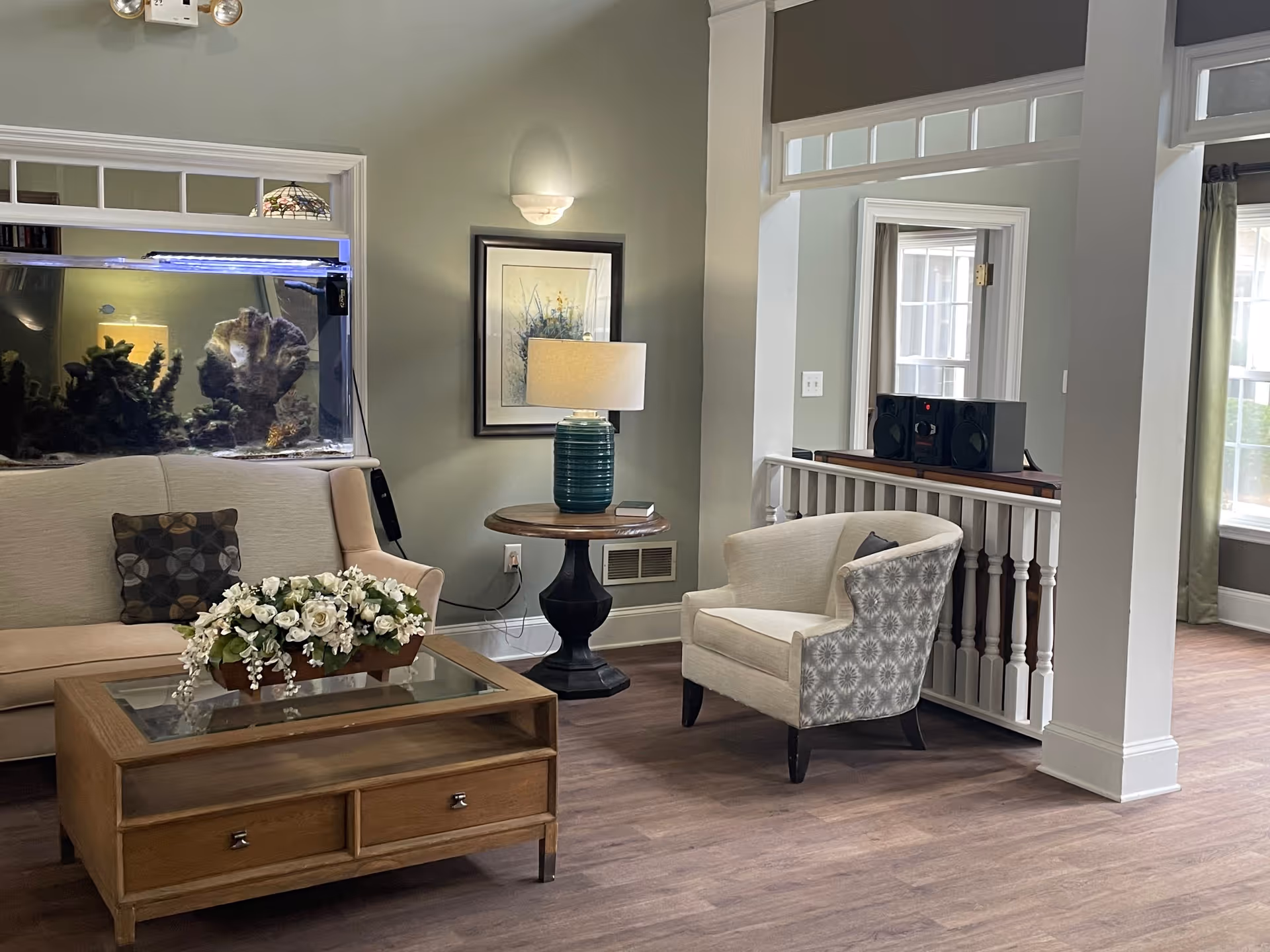 Bright common living room with a sofa, armchair, coffee table topped with a floral arrangement, side table with a lamp, and a large aquarium.