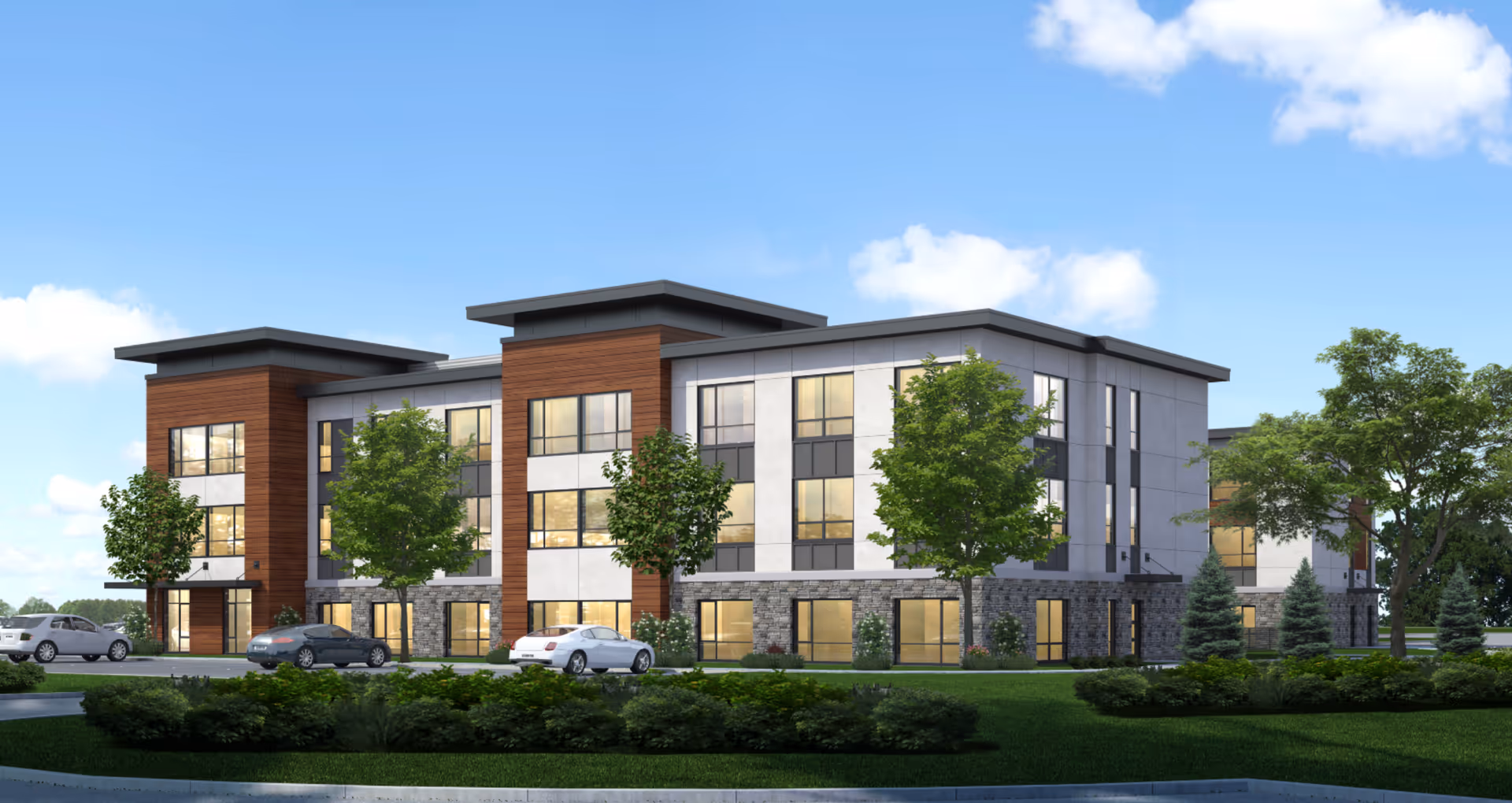 Exterior view of a modern three-story senior living facility building with large windows, stone and wood panel accents, surrounded by green trees and shrubs under a blue sky with some clouds. Several cars are parked in front of the building.