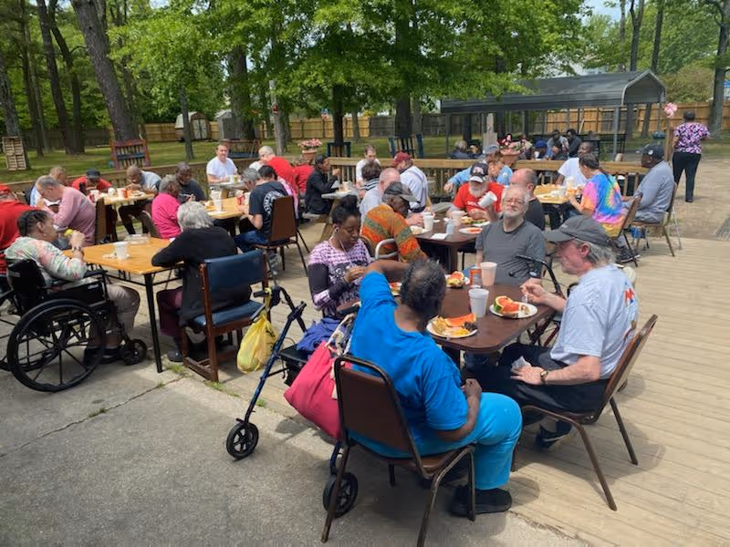 A group of elderly people sitting at outdoor tables enjoying a meal together in a shaded area with trees and a wooden fence in the background. Some individuals use wheelchairs and walkers, and the atmosphere appears social and relaxed.