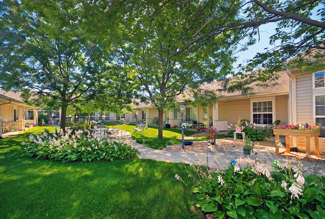 A sunny outdoor courtyard area with green grass, trees, and flower beds. There are several patio tables and chairs arranged along a paved walkway. Surrounding the courtyard are single-story beige buildings with windows and porches, decorated with potted plants and flowers.
