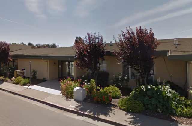 Single-story healthcare facility front with an entrance, flowering shrubs and small trees along the sidewalk.