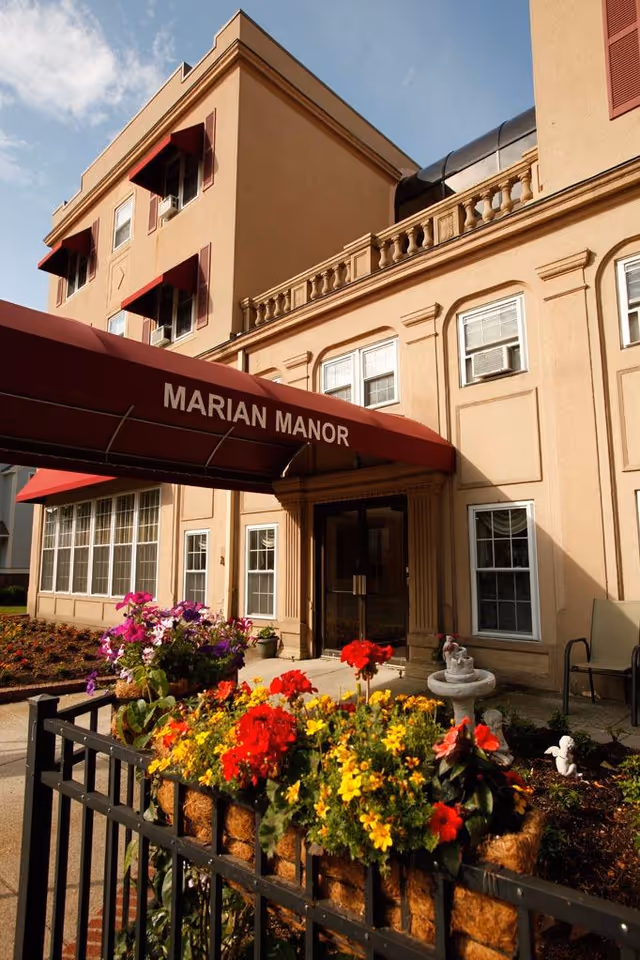 Entrance of Marian Manor building with a red awning displaying the facility name. The exterior is beige with multiple windows, and there is a garden area with colorful flowers and a small fountain in front.