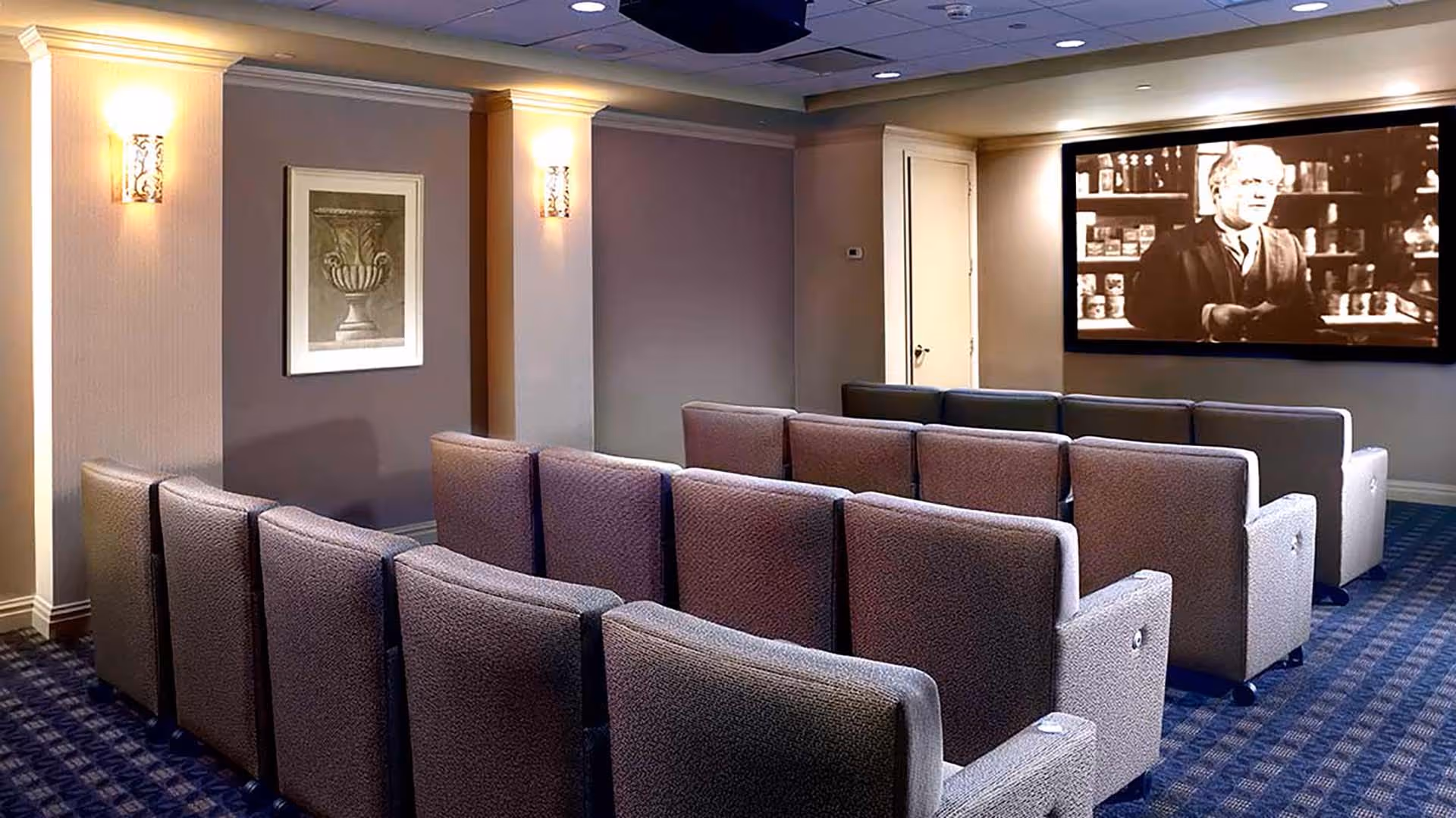 A small theater room with several rows of cushioned chairs facing a large screen showing a black and white movie. The room has soft lighting with wall sconces and a framed picture on the wall.
