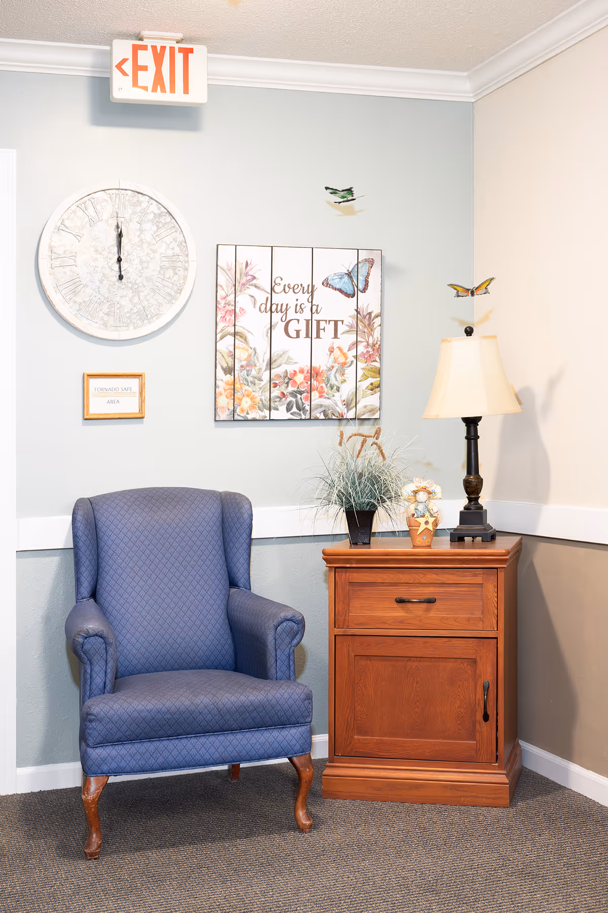 A cozy corner in a senior living facility featuring a blue upholstered armchair with wooden legs next to a wooden side table. The table holds a lamp with a beige shade, a small decorative plant, and a small figurine. On the wall above the chair is a large round clock, a sign that reads 'TORNADO SAFE AREA,' and a floral wall art piece with butterflies and the phrase 'Every day is a GIFT.' An illuminated exit sign is mounted near the ceiling.