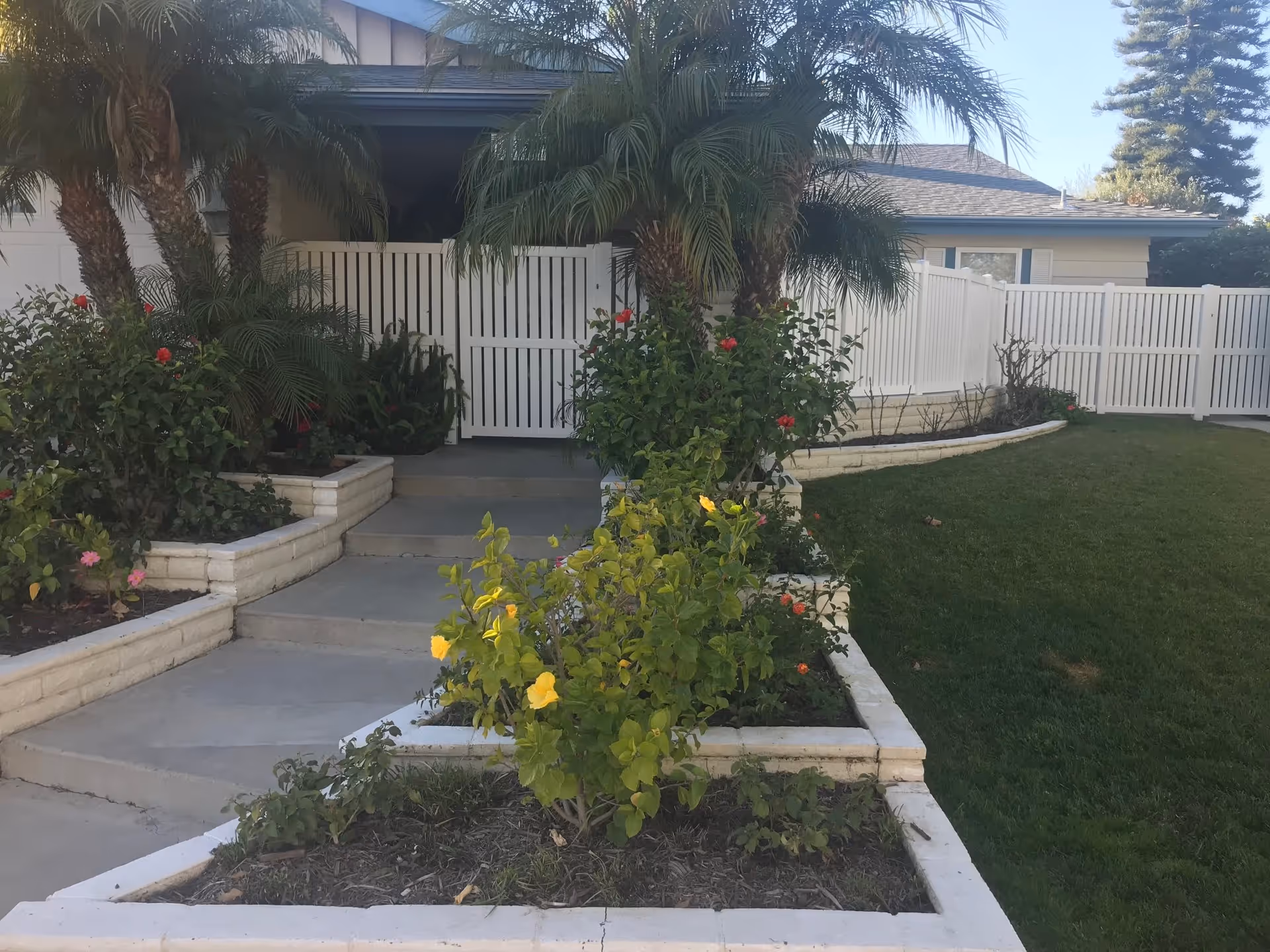 Front entrance of a single-story building with a white picket fence gate, surrounded by landscaped garden beds containing various green plants and colorful flowers, including yellow and red blooms. Palm trees are visible near the entrance, and a well-maintained lawn is on the right side.