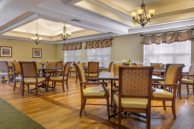 A well-lit dining room with multiple wooden tables and cushioned chairs arranged neatly. The room features large windows with blinds and valances, chandeliers hanging from a coffered ceiling, and framed artwork on the walls. The floor is wooden with a polished finish.