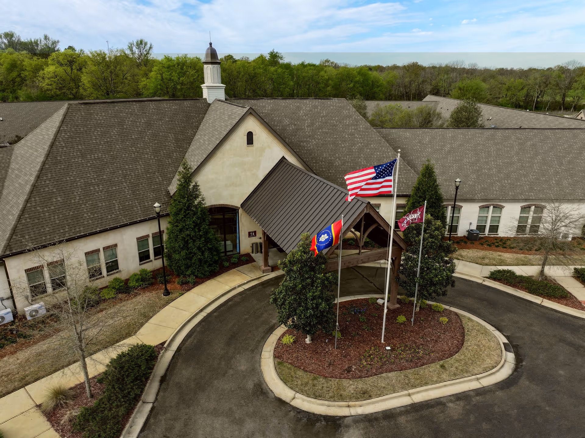 Aerial view of the entrance to The Goldton at Adelaide facility, showing a circular driveway with three flagpoles displaying the American flag, a state flag, and another flag. The building has a large roof with a small cupola and is surrounded by trees and landscaping.