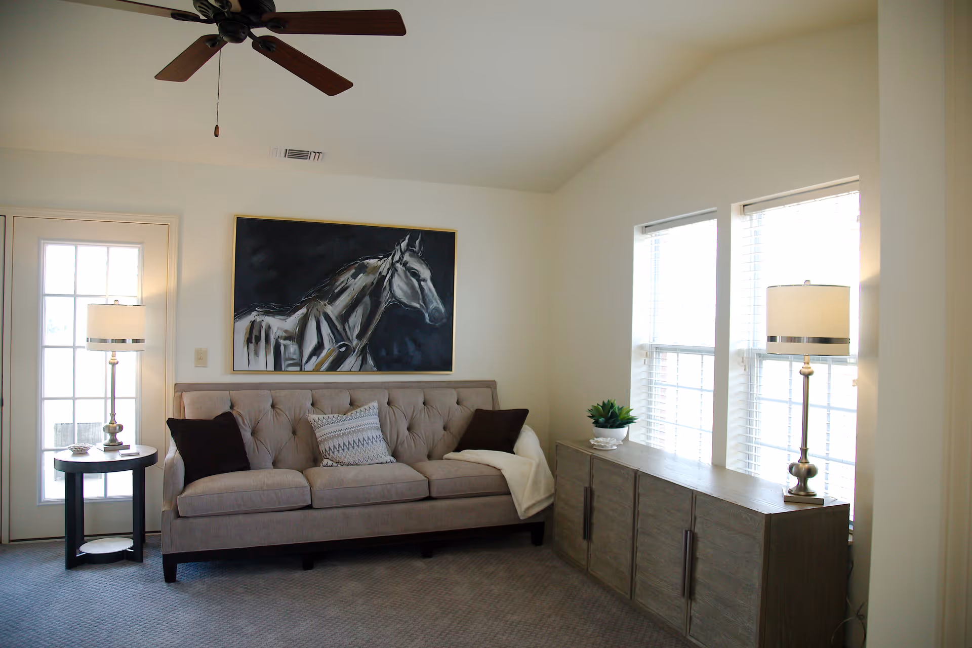 Bright living room with a tufted beige sofa beneath a large horse painting, flanked by table lamps, a sideboard and windows.