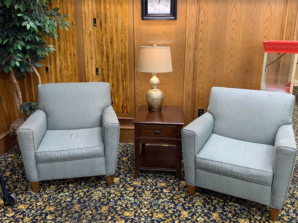 Two light blue armchairs with wooden legs are positioned on a patterned carpet with a dark wooden side table between them. On the table is a decorative lamp with a beige shade. Behind the chairs is a wood-paneled wall with a clock hanging above the lamp. A green leafy plant is partially visible on the left side, and a red popcorn machine is visible on the right side.
