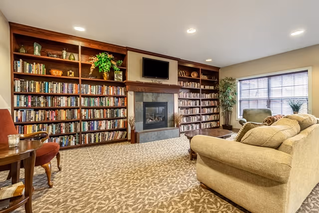 A cozy living room area with beige patterned carpet, a large beige sofa, and an armchair near a window with blinds. The room features built-in wooden bookshelves filled with books and decorative items, a fireplace with a mounted flat-screen TV above it, and a small wooden side table with a candle and a glass.
