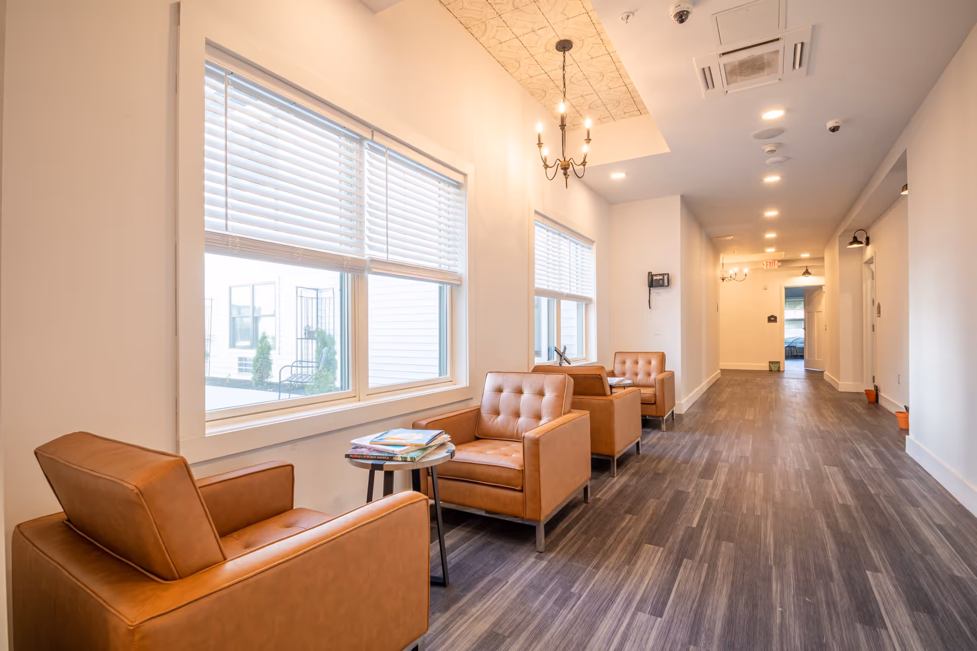A bright hallway with three brown leather armchairs lined up against a wall with large windows covered by white blinds. A small round table with magazines is placed between the first two chairs. The floor is covered with dark wood-like flooring, and the ceiling has recessed lighting and a decorative chandelier. The hallway extends into the distance with doors and light fixtures on the right side.