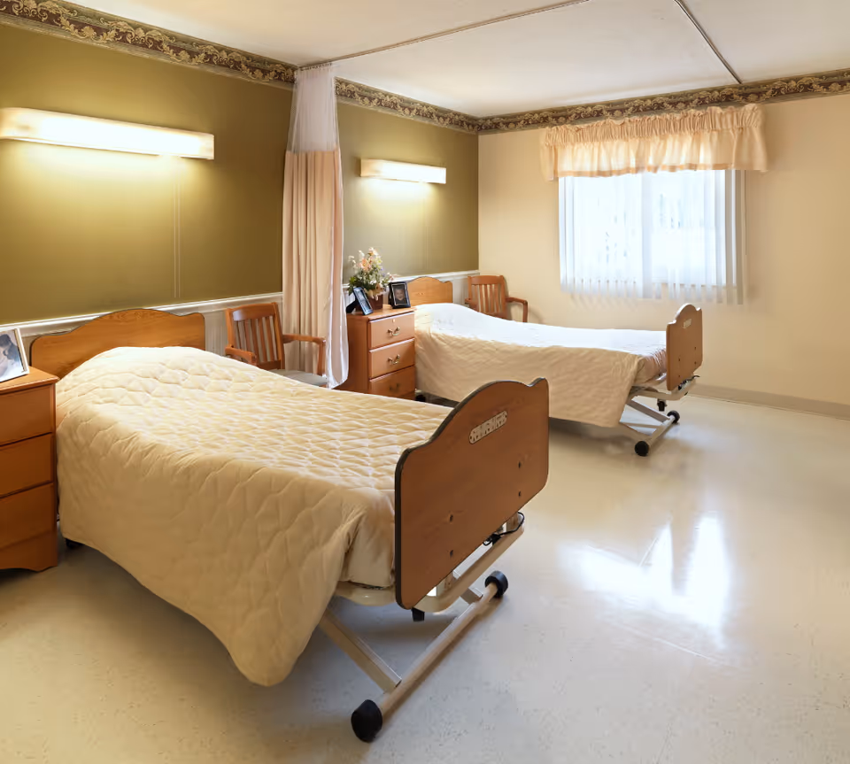 A clean and well-lit room with two hospital-style beds covered with beige quilts, wooden headboards, a wooden nightstand with framed photos and a flower arrangement, two wooden chairs, and a window with sheer curtains and a valance.
