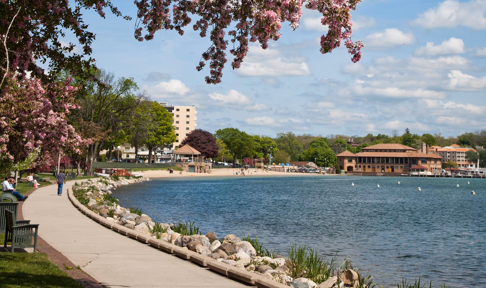 A scenic lakeside walkway lined with benches and flowering trees. People are walking and sitting along the path. Across the water, there are buildings and a sandy beach area under a partly cloudy sky.