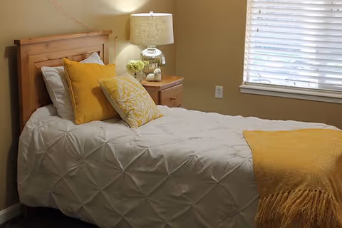 Small bedroom with a single bed dressed in white bedding and yellow pillows and throw, a wooden nightstand with a lamp, and a window with blinds.