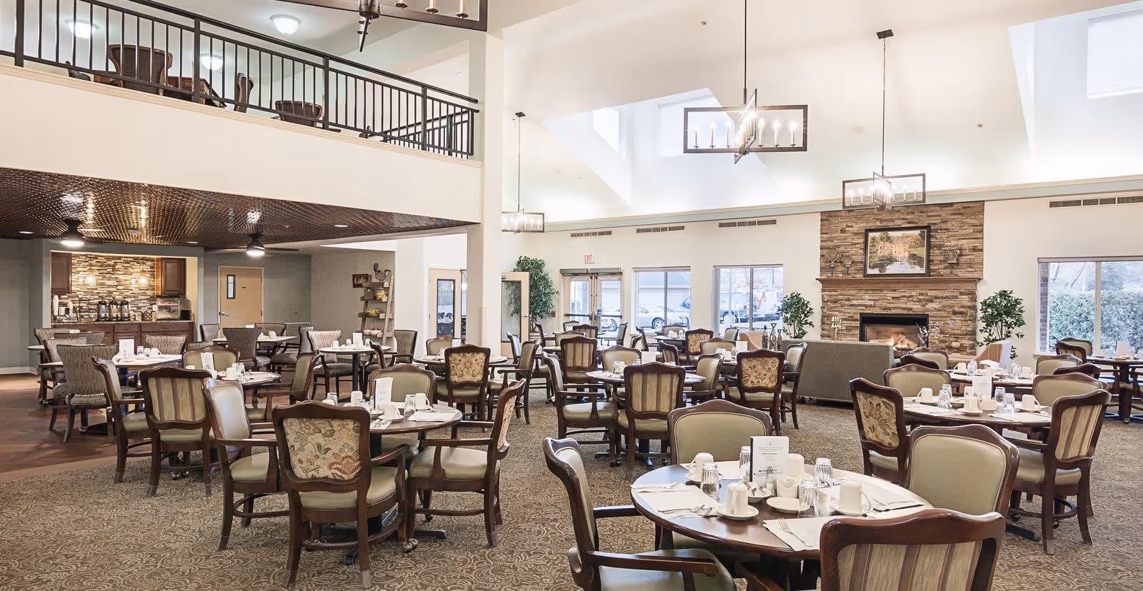 A spacious dining room in a senior living facility with multiple round tables set with cups, plates, and napkins. The room features upholstered chairs, a stone fireplace with a framed picture above it, large windows letting in natural light, and a mezzanine level with a railing. The ceiling has modern light fixtures and skylights.